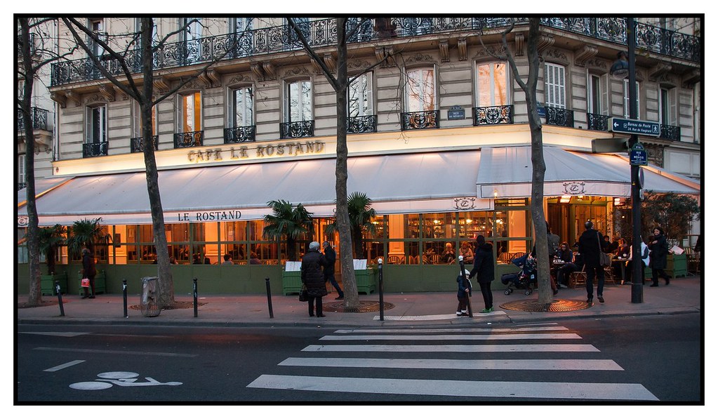 The illuminated terrace of Café Le Rostand at dusk. Parisian cafés