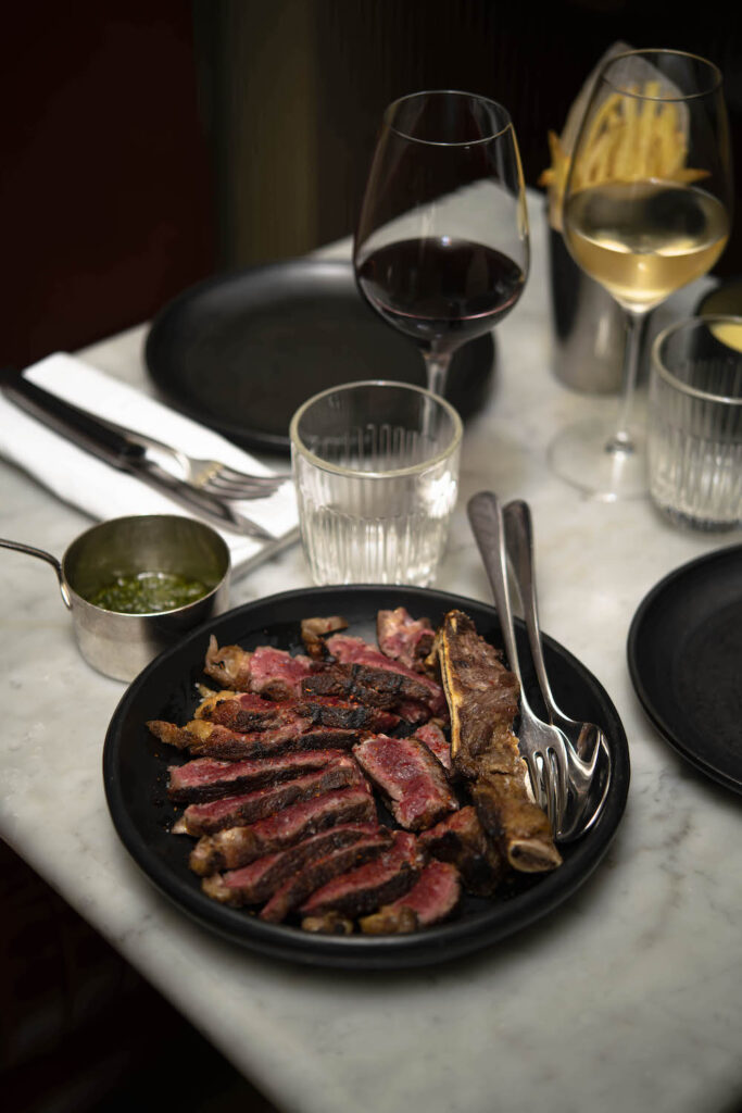 Grilled red meat dish with two glasses of wine at Aux Prés restaurant in Paris, showcasing Cyril Lignac’s mastery of French cuisine.