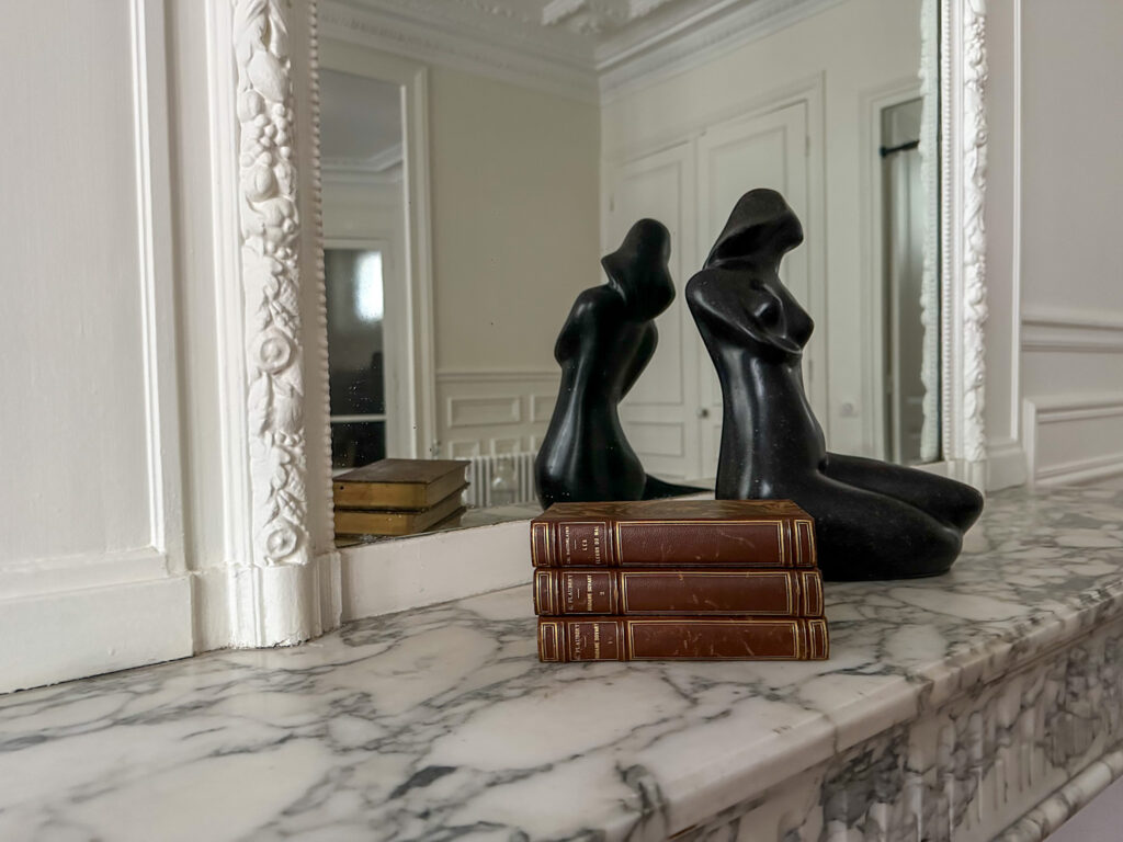 A stack of vintage leather-bound French novels placed on a white marble fireplace, accompanied by a black abstract sculpture, with a decorative mirror in the background.