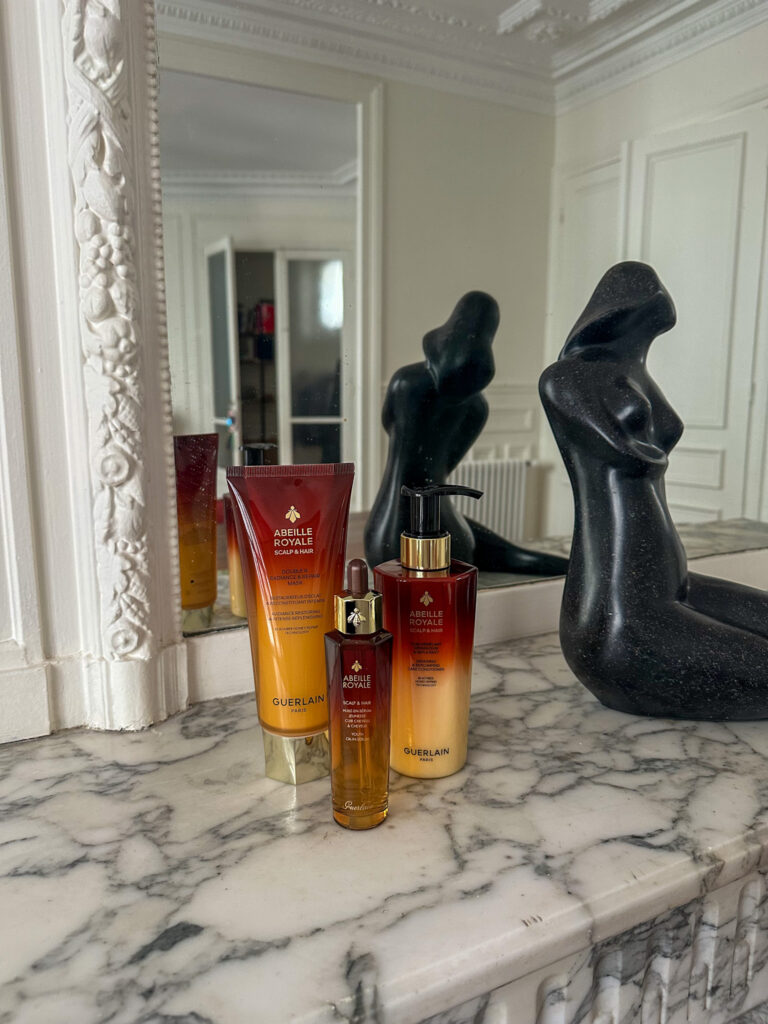 Guerlain Abeille Royale hair care collection displayed on a marble mantelpiece, featuring the shampoo, youth oil-in-serum, and repair mask.