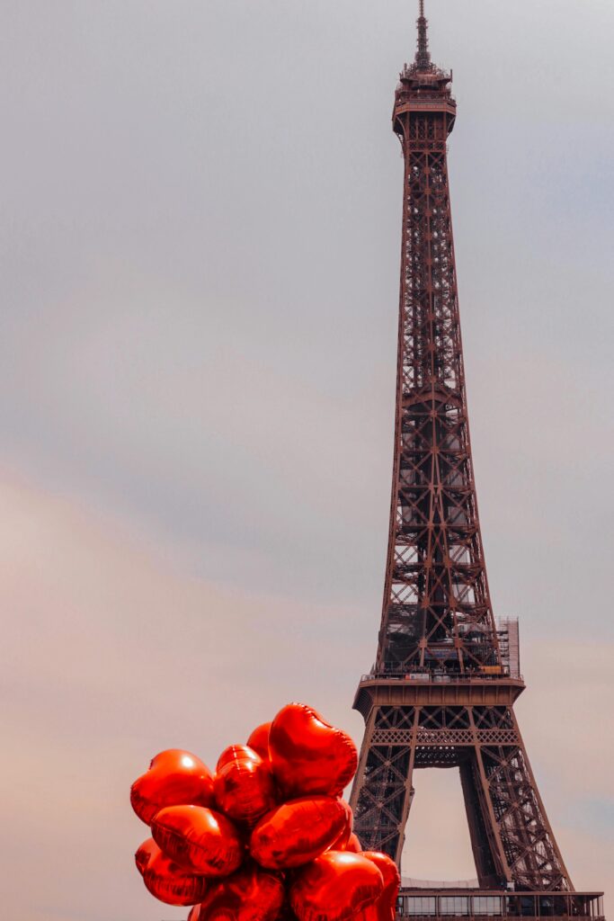 The Eiffel Tower with red heart-shaped balloons floating in the foreground against a soft gray sky.
