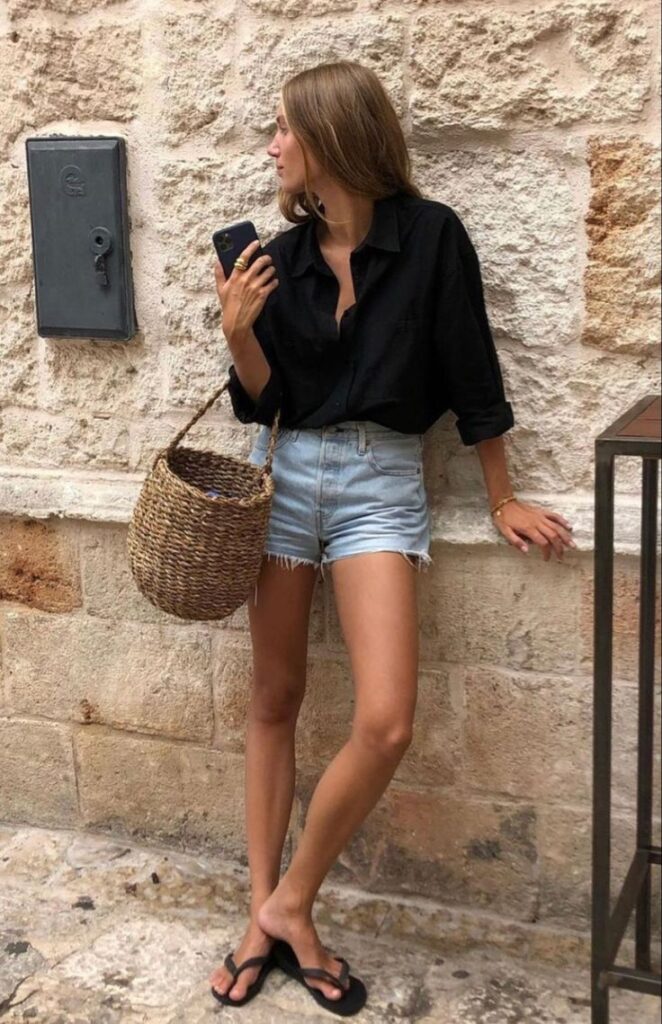 A woman in a black linen shirt and denim shorts carrying a round woven basket bag in a casual street setting.