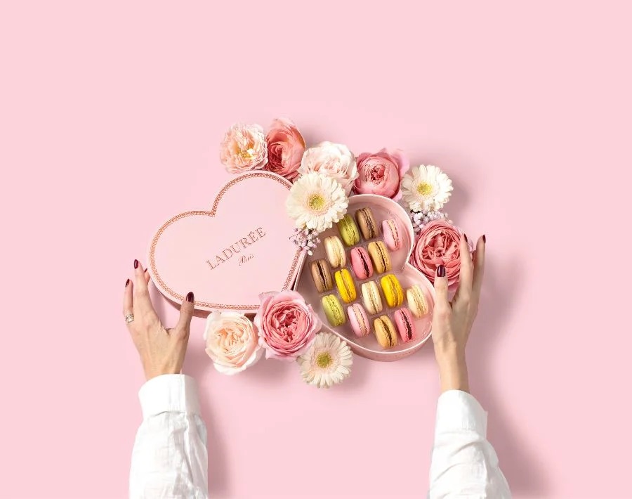 Ladurée heart-shaped box of macarons surrounded by pink and white flowers on a pastel pink background.