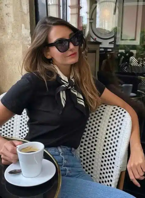 A woman enjoying coffee at a Parisian café, wearing a black and white silk scarf styled with a black t-shirt and jeans.