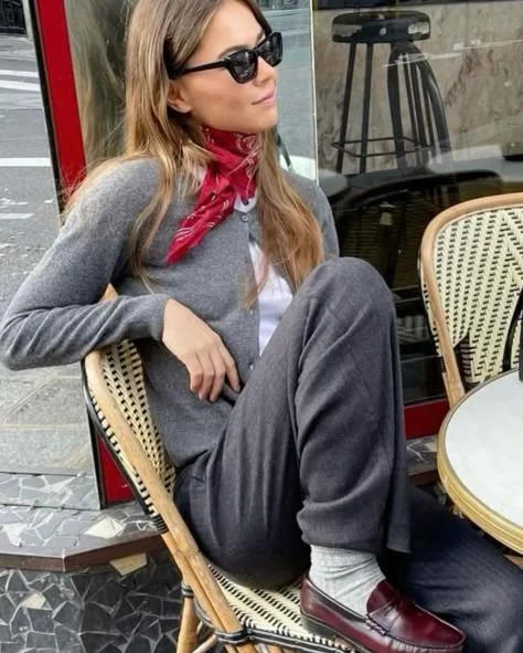 A woman wearing a red silk scarf tied around her neck, paired with a grey cardigan, trousers, and loafers in a Parisian café.