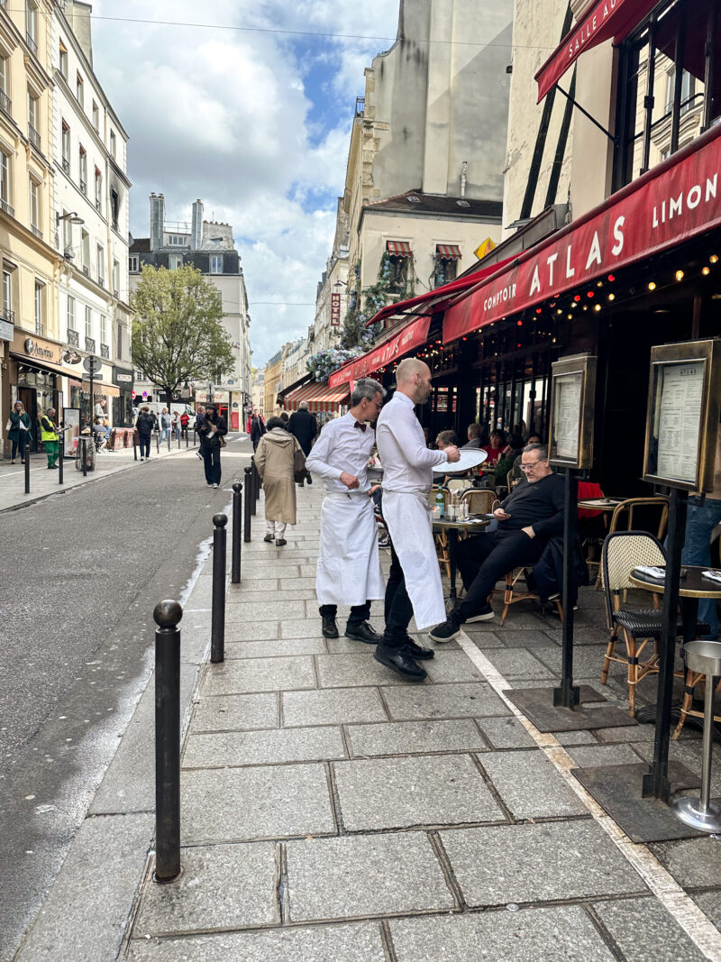 Two waiters in white aprons serving guests outside a classic Parisian café with red awnings in early spring