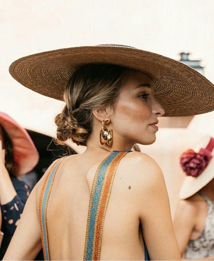 Woman in a backless dress wearing a wide-brimmed straw hat and sculptural gold earrings at a wedding