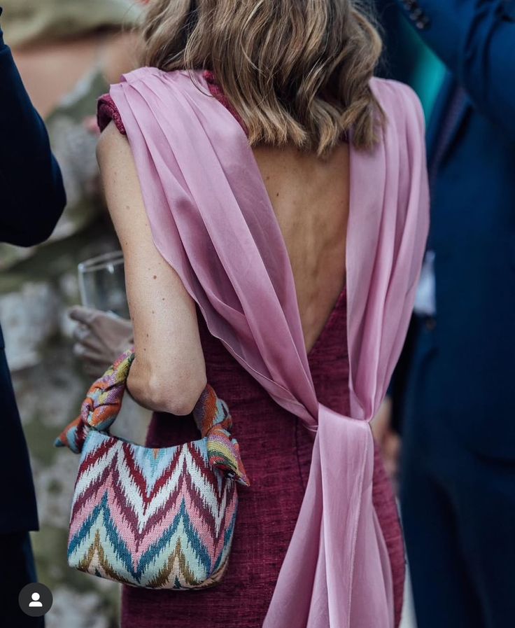 Woman wearing a burgundy backless dress with a flowing pink cape and a colorful woven handbag