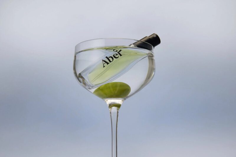 Abel mini perfume bottle floating inside a coupe glass with a green olive, shot against a minimal backdrop