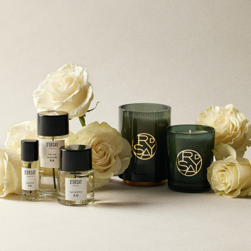 Maison D’Orsay perfume bottles and candles surrounded by cream-colored roses on a neutral background