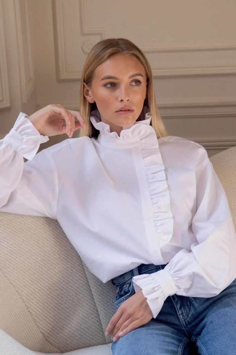 Woman seated wearing a crisp white Rosaé Paris blouse with ruffled details
