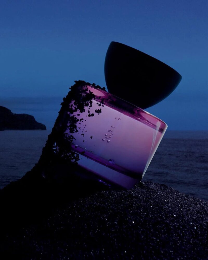 Vyrao Witchy Woo purple perfume bottle with black cap resting on volcanic sand at dusk by the sea