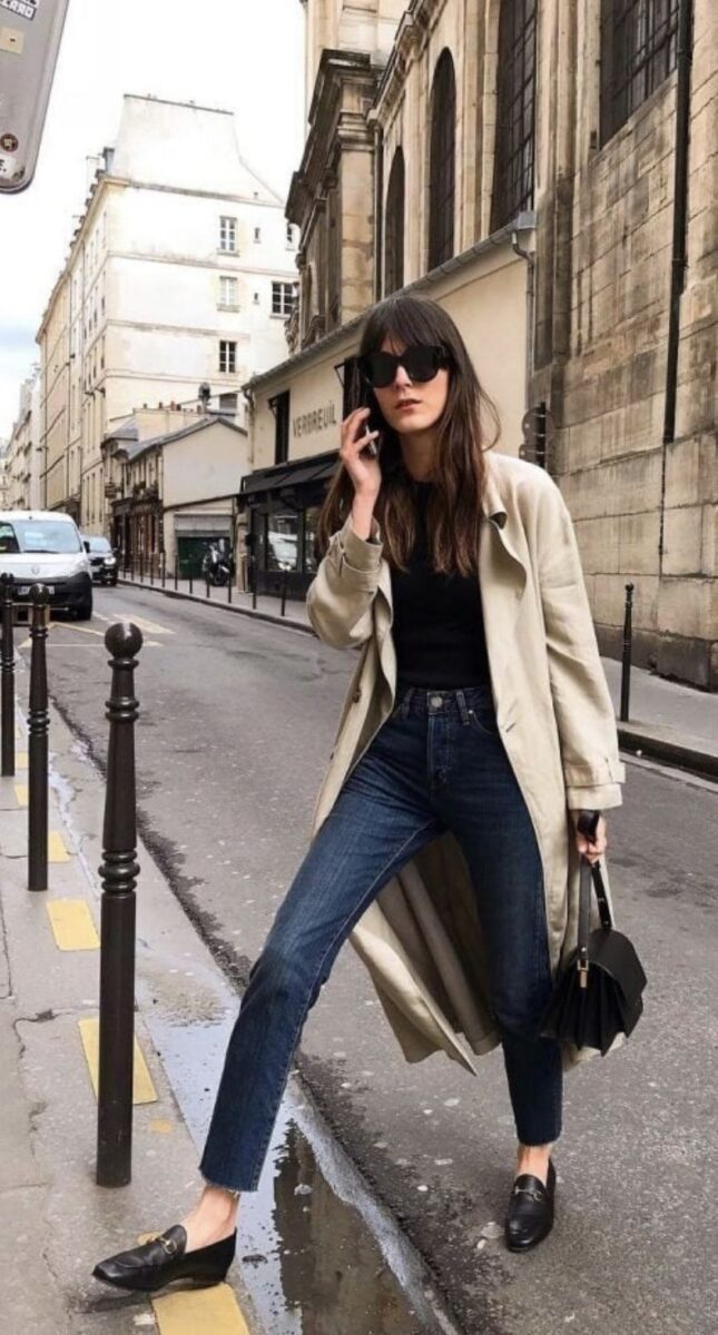 Woman walking in Le Marais in Paris wearing dark wash straight jeans, loafers, a trench coat and sunglasses, the epitome of Parisian style.