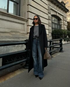 Woman wearing a long black coat, grey sweater, wide-leg jeans, and pointed boots – a chic street style outfit for what to wear in Paris in the fall.
