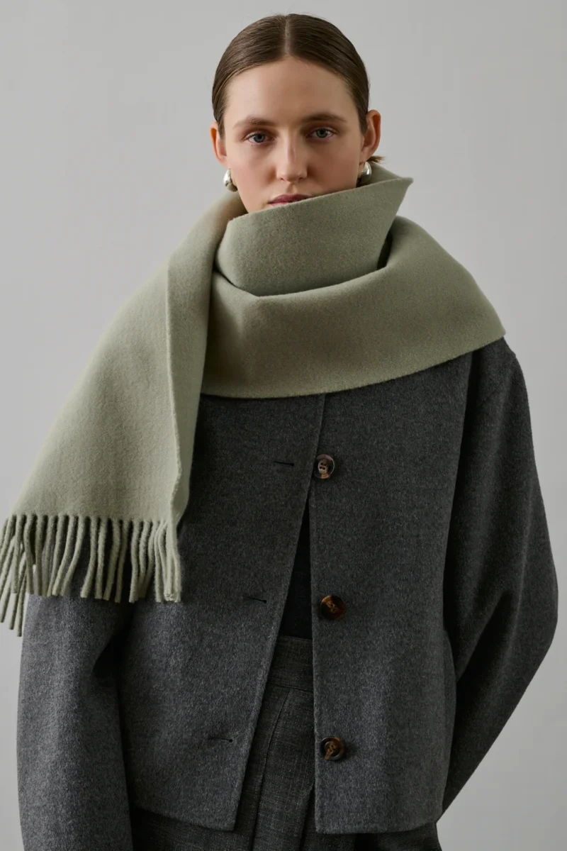 Model wearing a French scarf by Soeur, pale green wool scarf with fringe, wrapped around the neck.