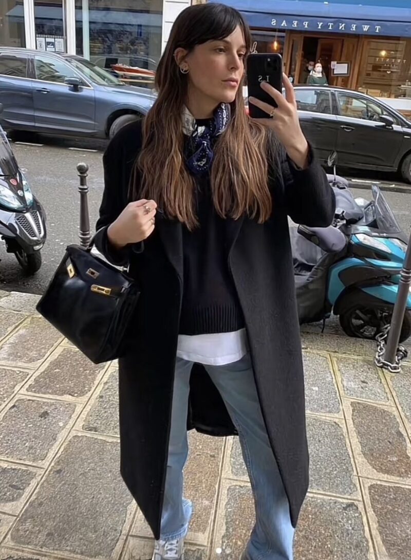 Woman wearing a long black coat, layered knit, silk scarf, light-wash denim, and sneakers — casual winter street style in Paris.