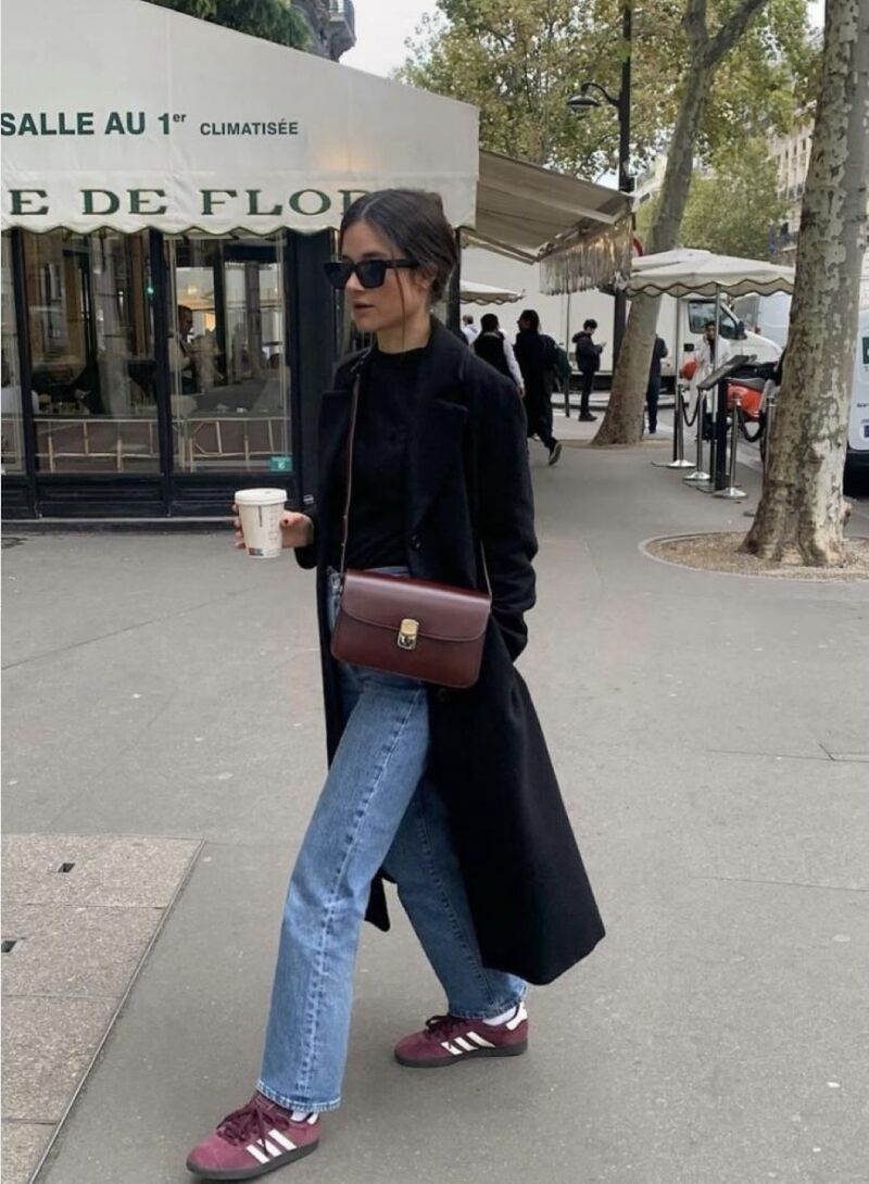 Woman wearing a long black coat, straight-leg jeans, burgundy Adidas sneakers, and a crossbody bag — casual Paris winter outfit.