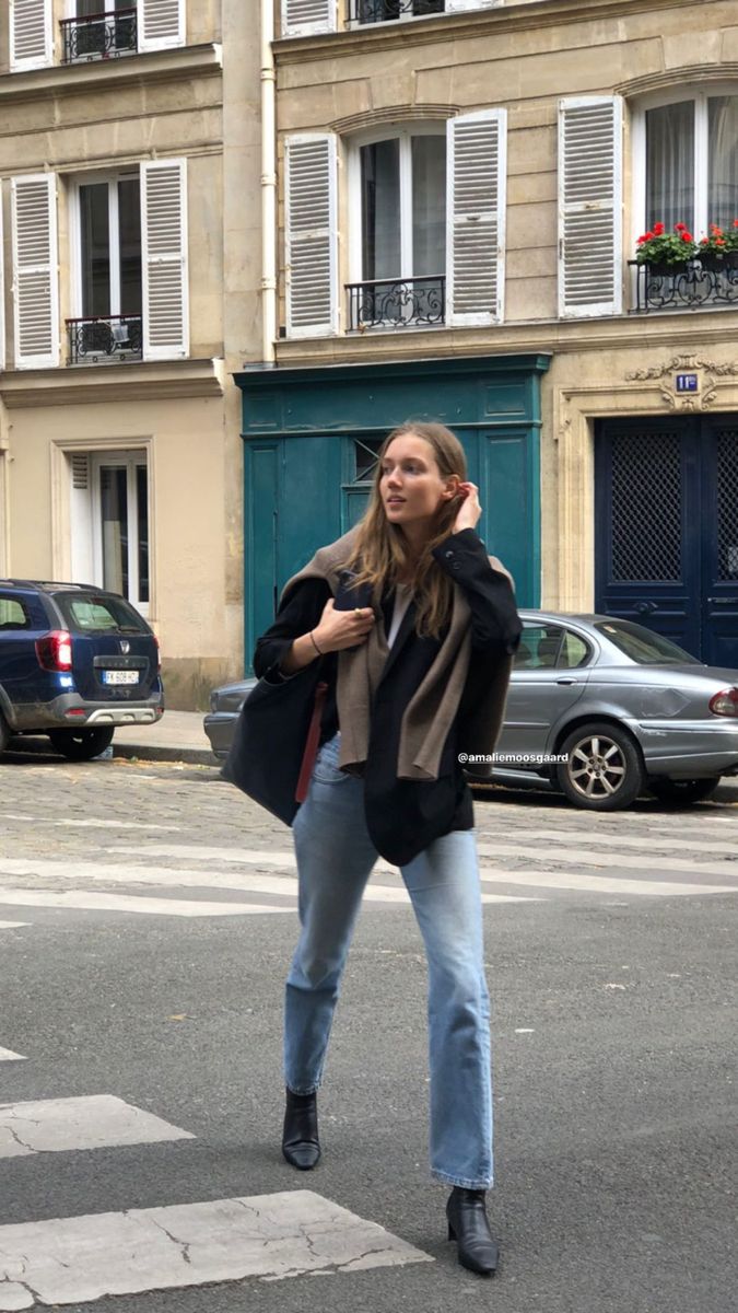 Woman wearing a black blazer, knit sweater layered over shoulders, straight denim, and black ankle boots — layered Paris winter styling.