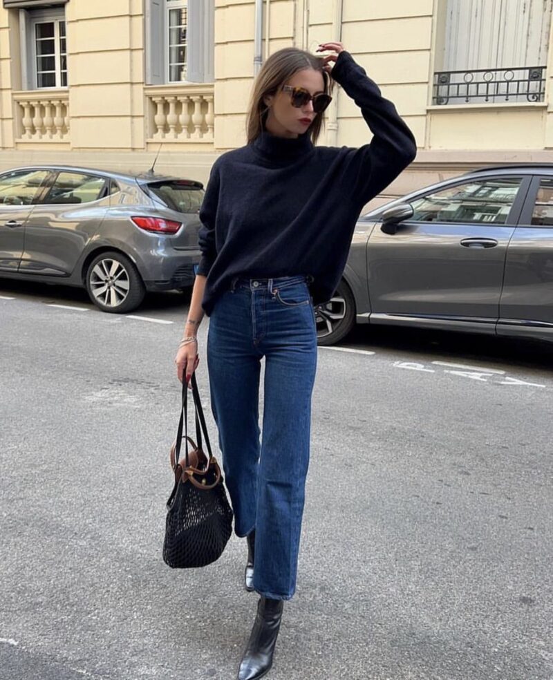 Woman wearing a navy turtleneck sweater, dark straight jeans, black ankle boots, and a bucket-style tote — minimalist Paris winter outfit.