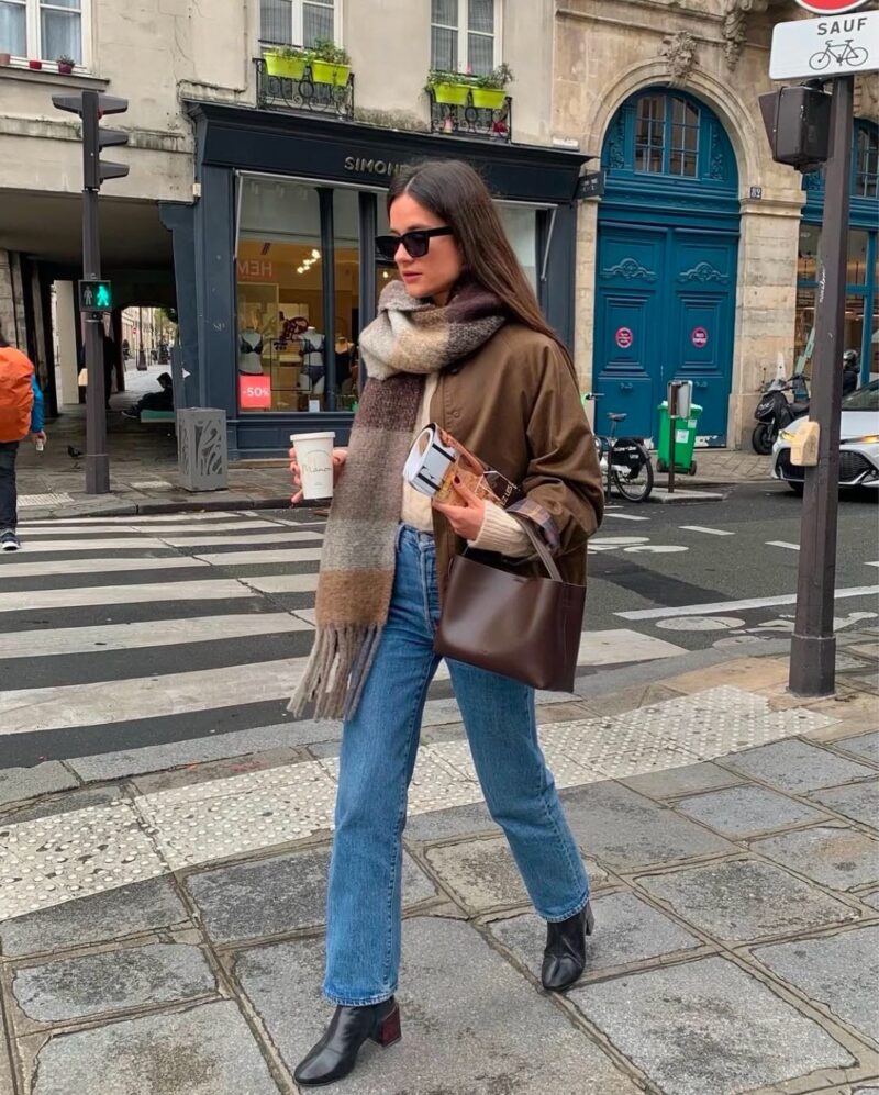 Woman wearing a cozy oversized scarf, brown jacket, straight-leg jeans, and black ankle boots — casual Paris winter look.